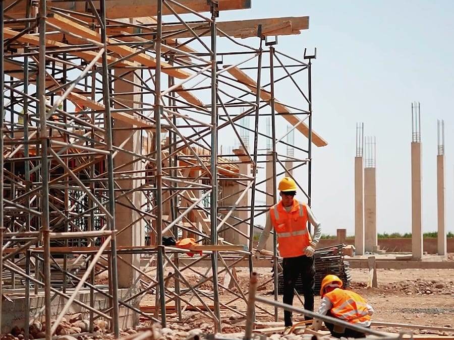Chantier en août 2026 : échafaudages, coffrages bois et ferraillage au premier plan.