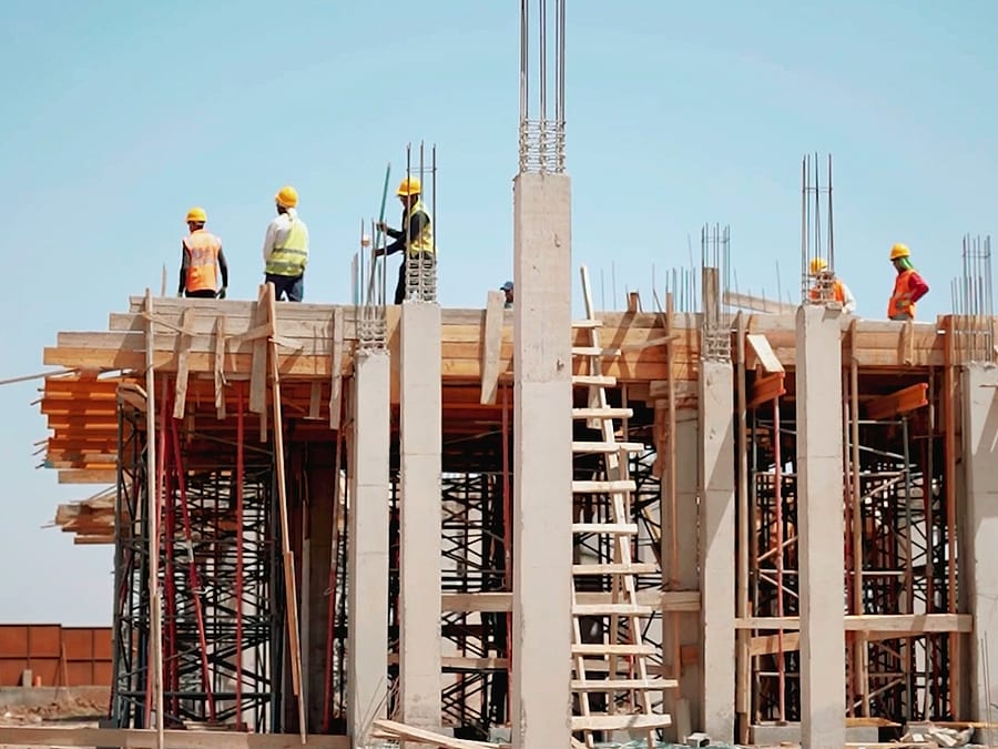 Chantier en août 2026 : ouvriers sur une dalle en cours de coffrage, poteaux et ferraillage.